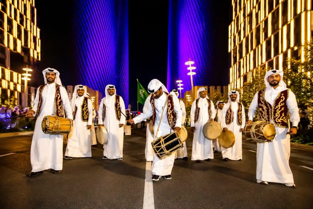 Traditional Gulf drummers and percussion performers in white kanduras performing a cultural procession at a luxury evening event, surrounded by illuminated skyscrapers and vibrant city lights.