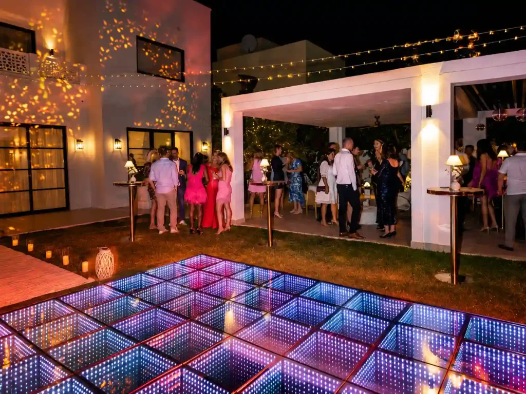 Luxury outdoor event featuring an illuminated LED infinity dance floor, elegant villa setting, festoon lighting, and guests socialising during an evening celebration.