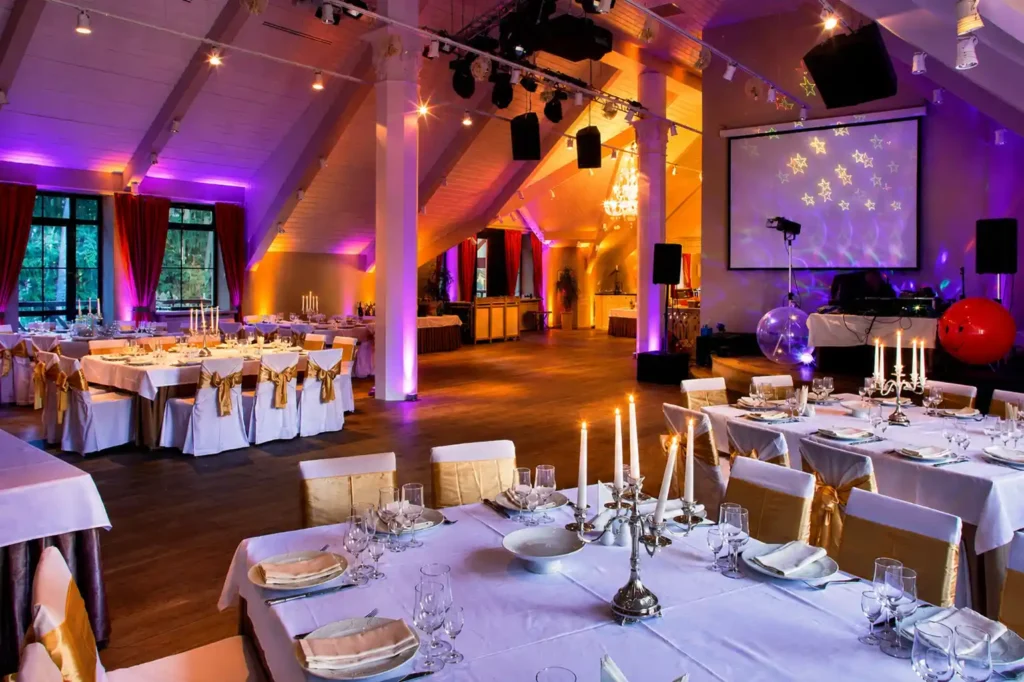Elegant event venue with purple and amber uplighting, banquet tables, DJ setup, projection screen and professional event lighting creating a luxury atmosphere for a corporate event or wedding.