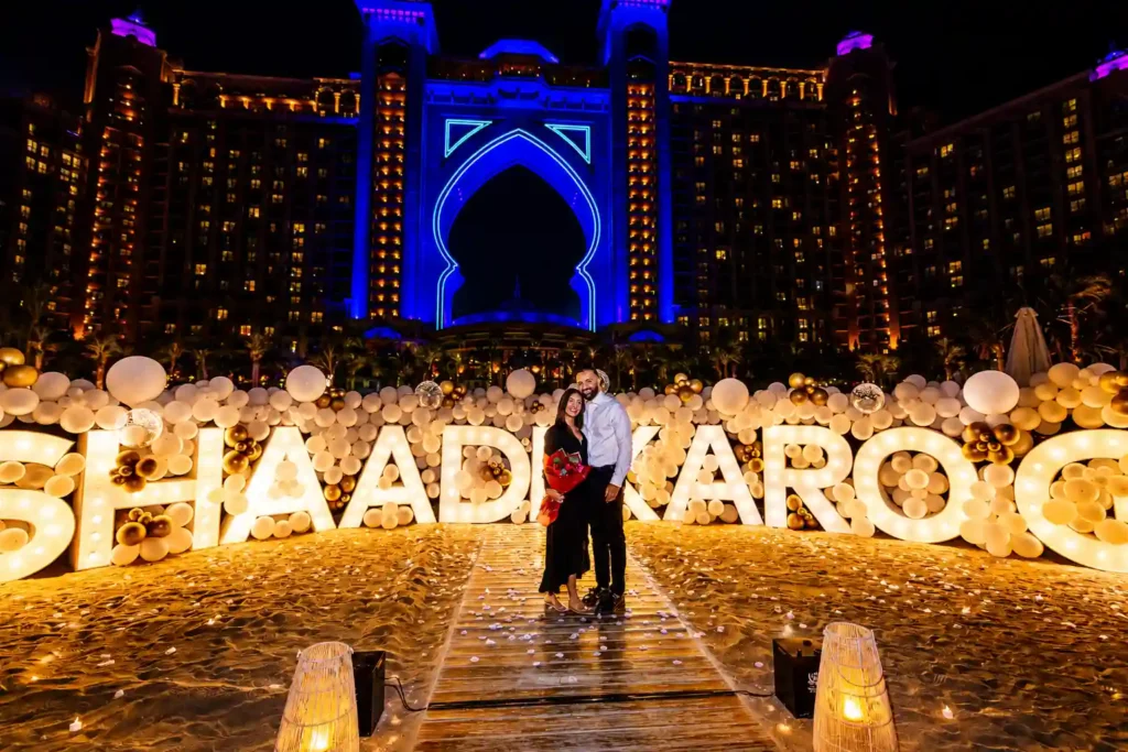 **Alt Text:**Romantic beach marriage proposal in Dubai featuring illuminated LED name letters, elegant white and gold balloon décor, a couple celebrating on a wooden walkway, and the iconic Atlantis The Palm illuminated in the background at night.
