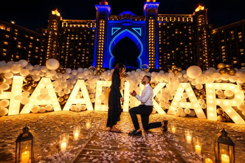 Romantic beachfront proposal at night in Dubai featuring a man on one knee proposing with an engagement ring, surrounded by illuminated giant "SHAADI KAROO" letters, white and gold balloon decorations, rose petals, and the iconic Atlantis The Palm illuminated in the background.