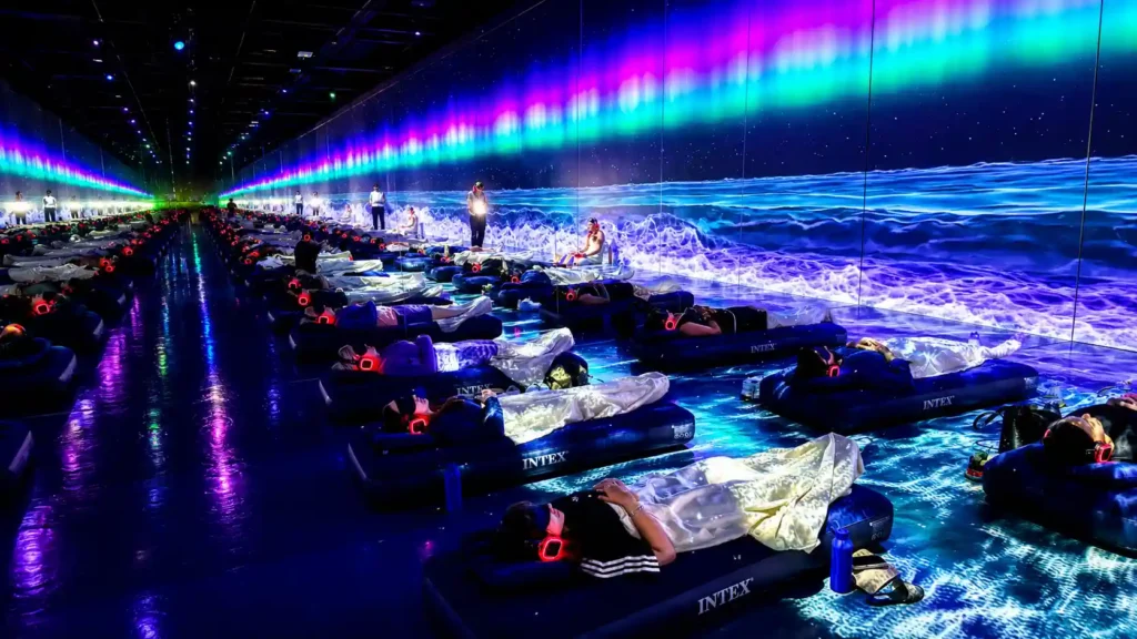 People relaxing on inflatable loungers with wireless headphones inside an immersive digital art exhibition featuring vibrant ocean waves and aurora projections across large LED walls, creating a tranquil audiovisual experience.
