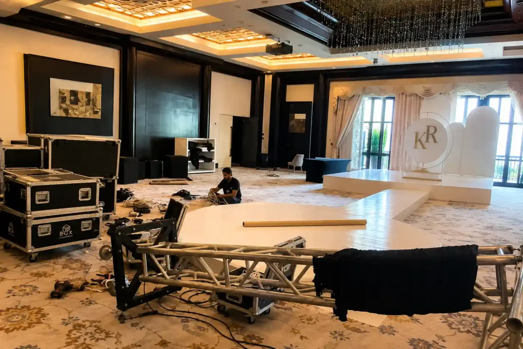 Behind the scenes of a luxury event production in Dubai, with technicians preparing a white stage and runway, organising cables, trussing, and AV equipment inside an elegant ballroom before the event begins.
