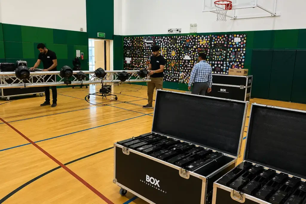Behind the scenes of a school event production with technicians assembling stage lighting on aluminium trussing, flight cases of lighting equipment, and AV gear inside an indoor sports hall before the event begins.
