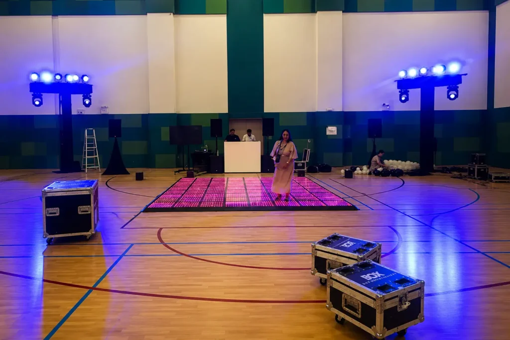 Behind the scenes of a school event setup featuring an illuminated LED dance floor, professional DJ booth, stage lighting towers, flight cases, and AV equipment inside a large indoor sports hall before guests arrive.
