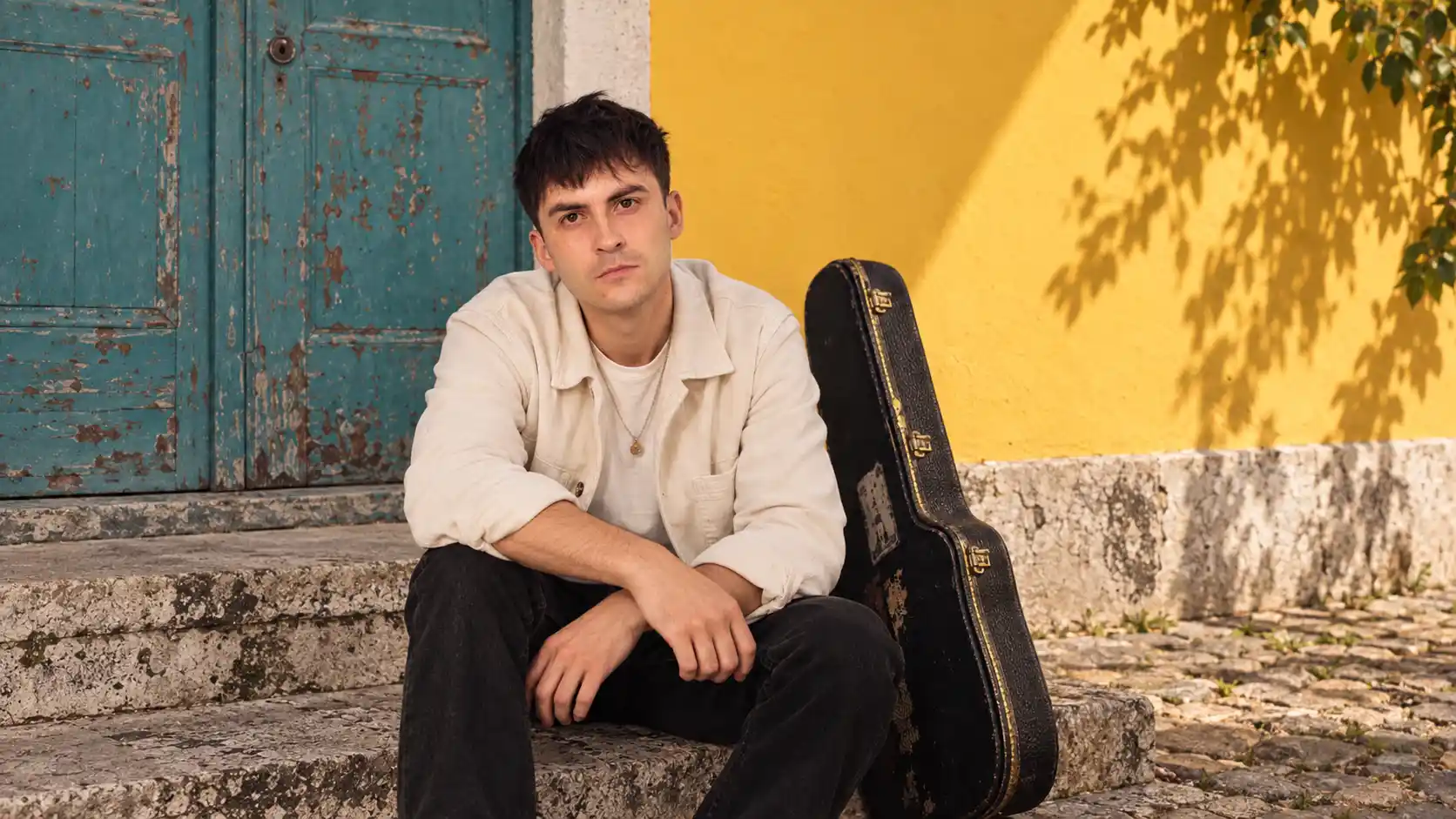 Greg Pearson seated beside a guitar case against a colourful turquoise door and yellow wall.