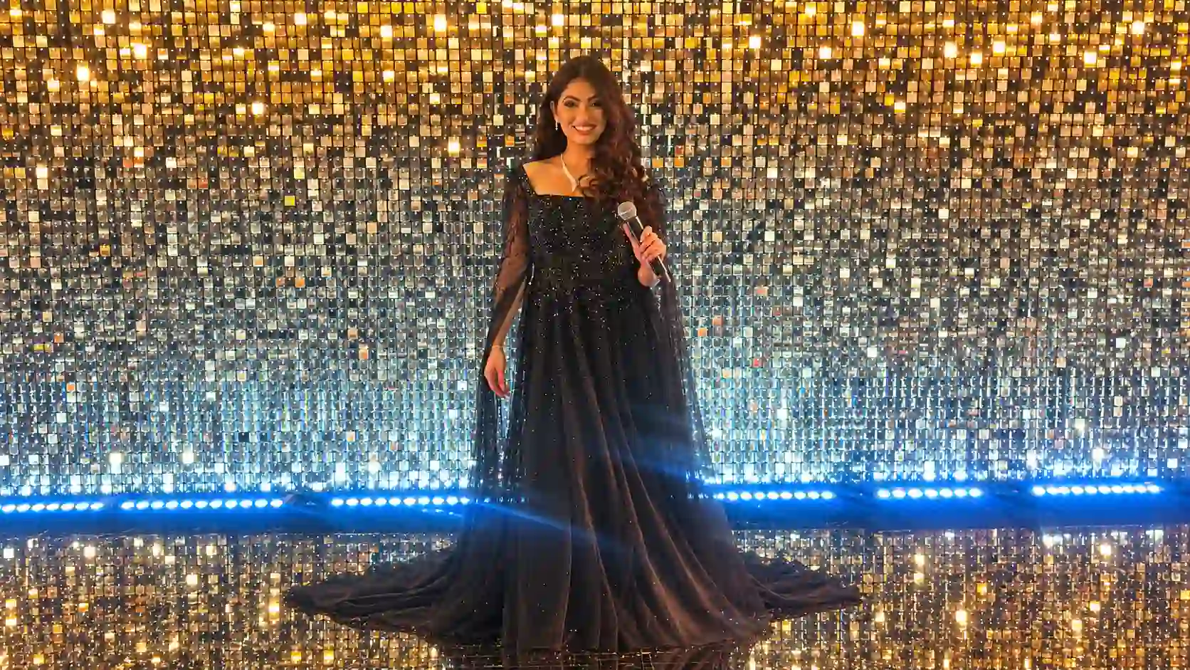 Joelle holding a microphone in a black evening gown against a shimmering gold and silver backdrop with blue lighting.