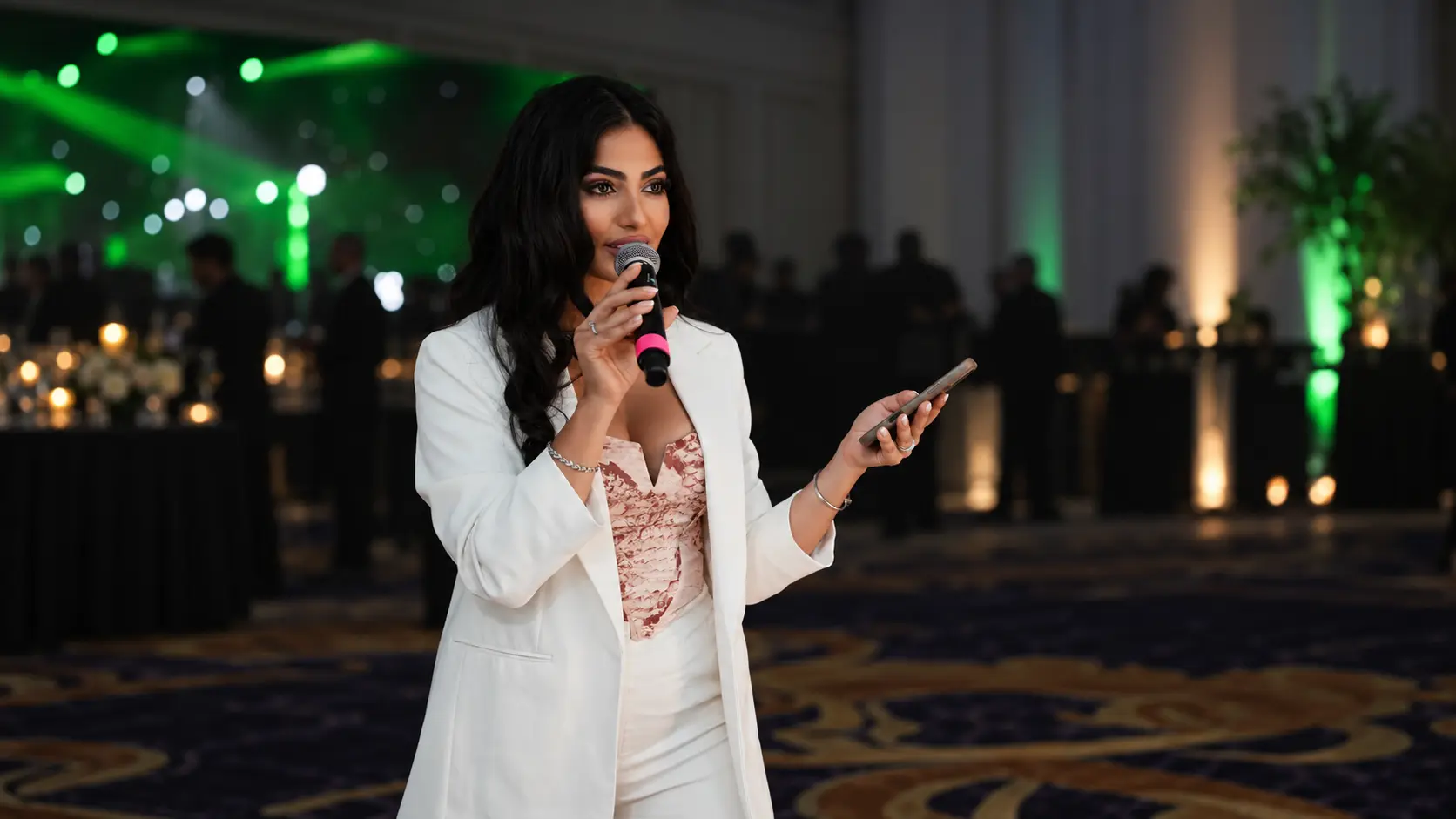 Joelle speaking into a microphone and holding a phone, wearing a white suit at an indoor event with green and gold lighting.