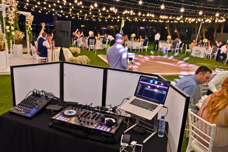 Wedding DJ booth with controller, mixer and laptop overlooking an outdoor dance floor, with a saxophonist and guests beneath warm fairy lights.