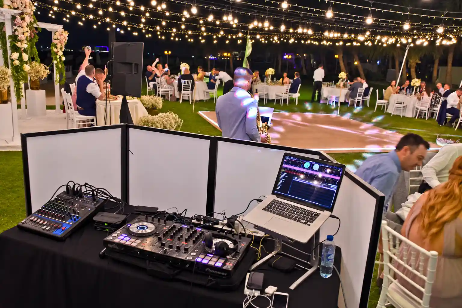 Wedding DJ booth with controller, mixer and laptop overlooking an outdoor dance floor, with a saxophonist and guests beneath warm fairy lights.