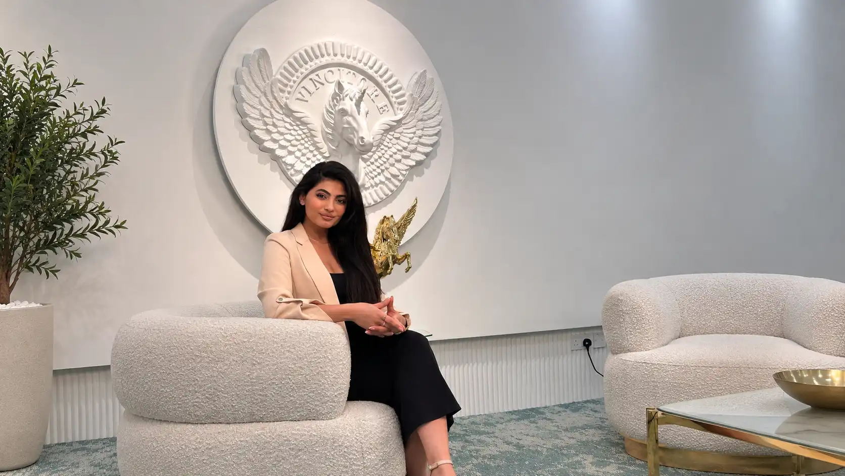 Joelle seated in a cream armchair, wearing a beige blazer over a black outfit beneath the Vincitore winged horse emblem.